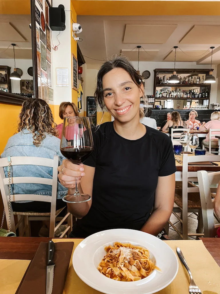 A woman smiling to camera, holding a wine glass on one hand. There's a plate of pasta with sauce in front of her