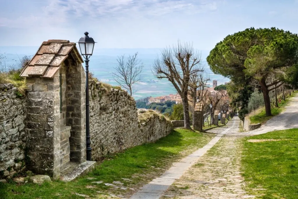 An image of Cortona, with the ruins of a stone wall on the left, and a path lined with trees