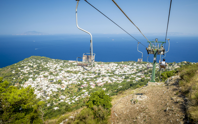 chairlifts over a white city that stands out in the middle of lush green vegetation, and the sea in the distance