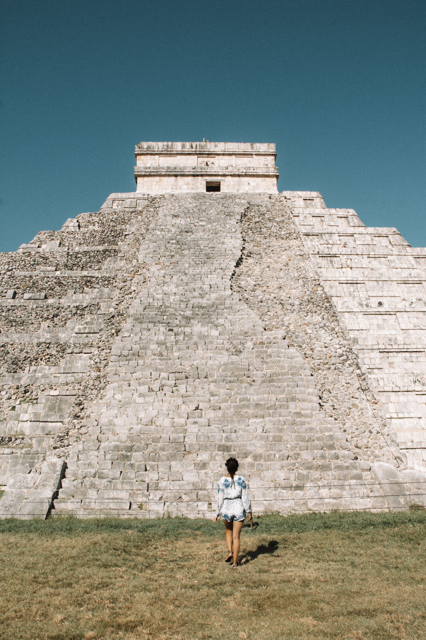 Woman with a white and blue playsuit standing right in front of the biggest pyramid in Chichen Itza.