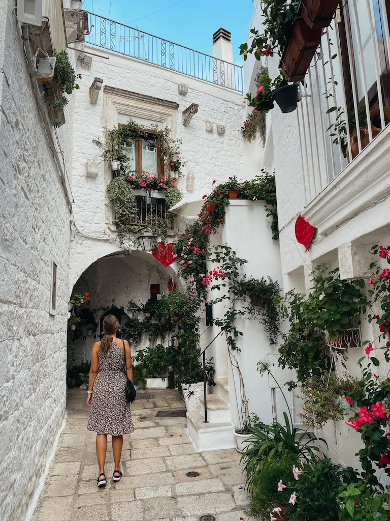 A woman standing in a narrow alley in Cisternino, with brimming flower pots hanging from white facades to her right