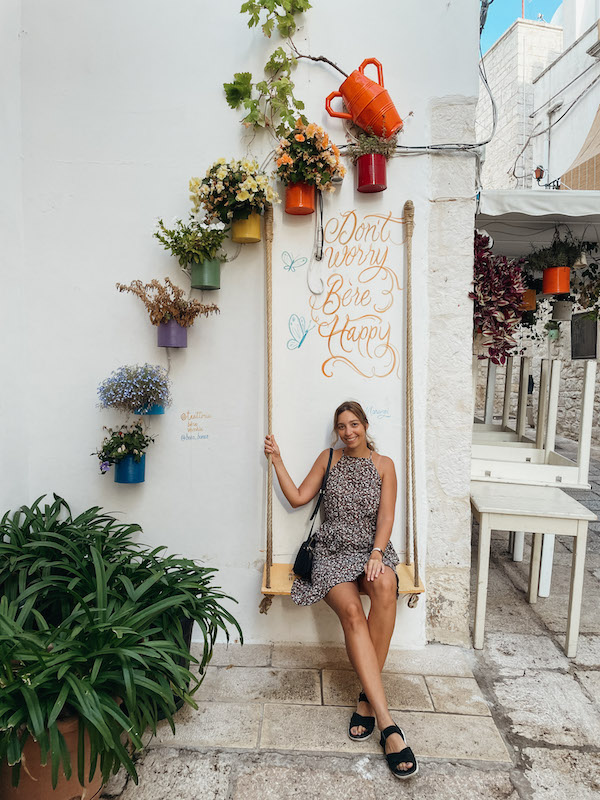 A woman sitting on a swing located next to a white wall, with a sign saying ''Don't worry, be happy'' behind her