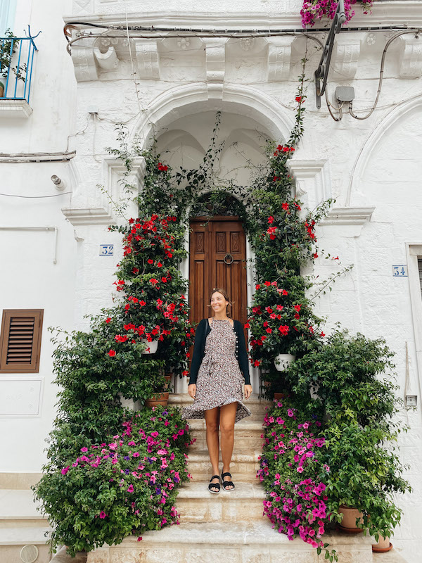 A woman standing on a flight of stairs in front of a door, with brimming flower pots on both sides of her