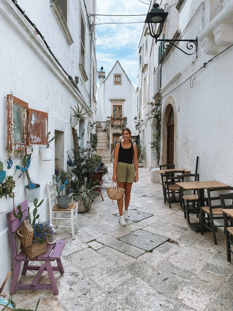 A woman wearing a black tank top and green shorts, standing in a narrow street lined by café tables and plants