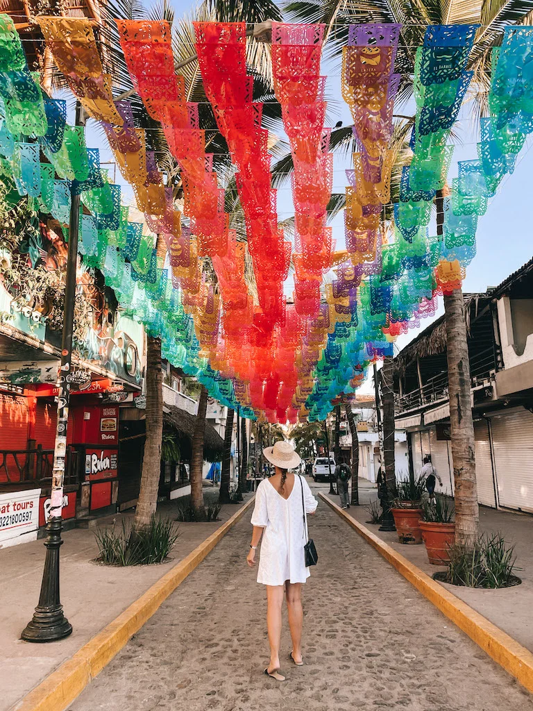 Woman with a white dress and straw hat standing under a series of colorful  paper flags.