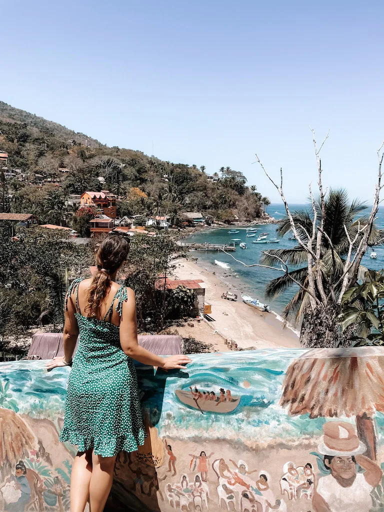 Woman in a green polka dot dress standing in front of a mural and looking down towards a bay.