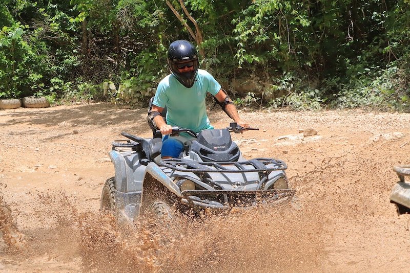 A man wearing a helmet and a blue shirt is riding an ATV through a muddy trail.