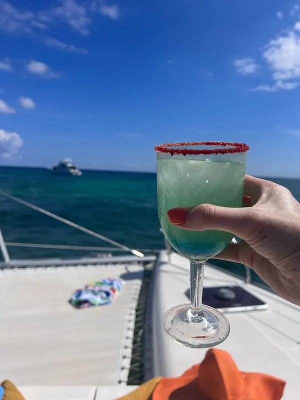 A hand with red nails holding a red-rimmed glass witha. green cocktails, a boat in the background.