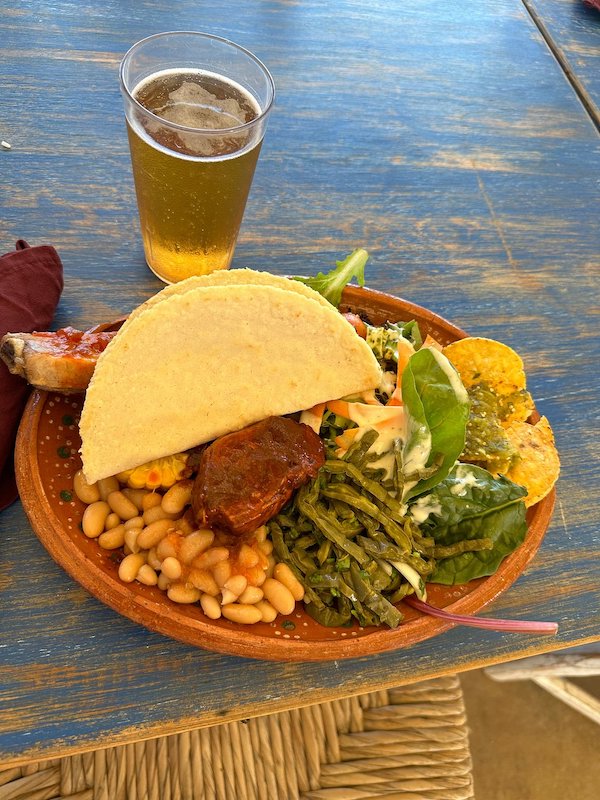 A brow clay plate filled with veggies, beans, tortillas and a beer in the background.