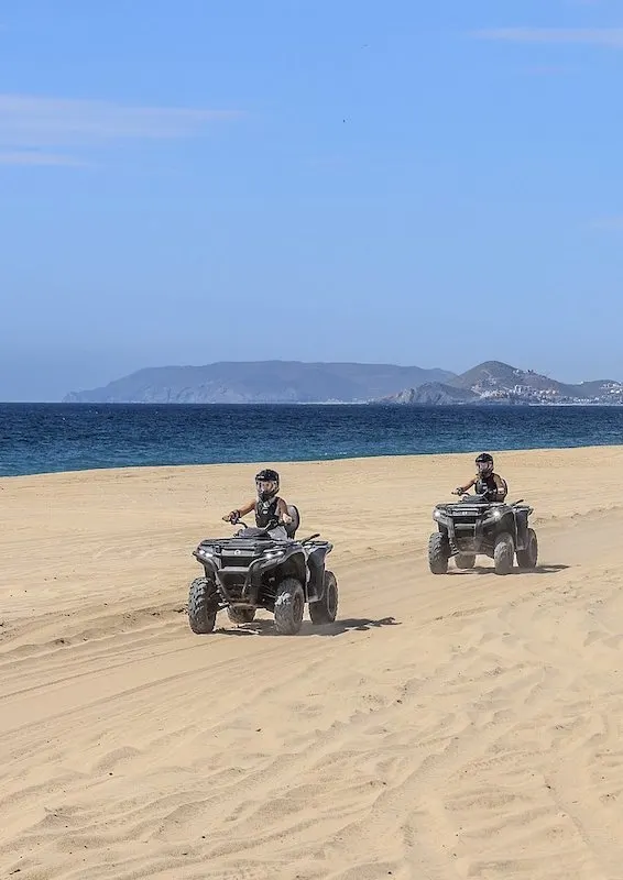 Two riders on black ATVs wearing a helmet and riding on the beach with the ocean in the background during an ATV tour in Cabo.