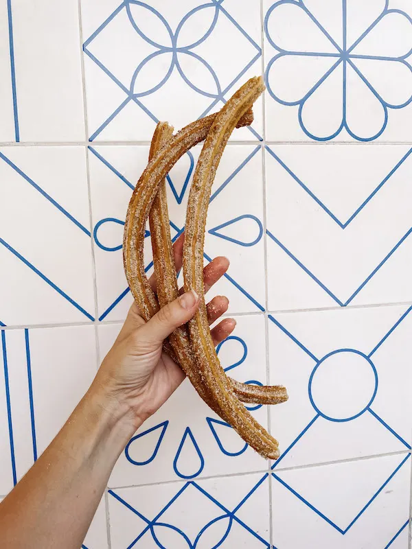 A hand holding 3 churros in front of a white and blue wall.