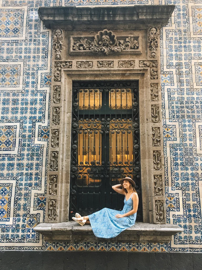 A woman wearing a blue dress and a brown hat is sitting on a windowsill in Mexico City, in a building covered in azulejos.