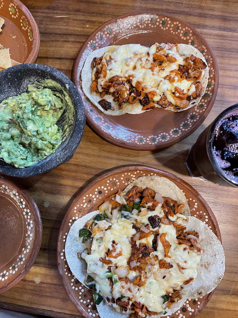 Multiple clay plates on a table with tacos covered with cheese and meat, and a guacamole bowl.