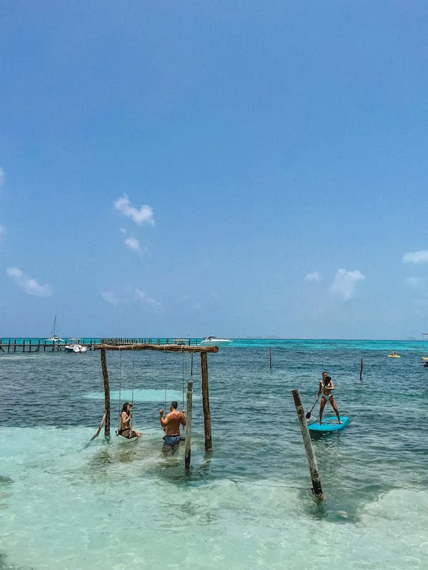 Two people on a swing in the water and a woman paddleboarding at a beach club in Isla Mujeres.