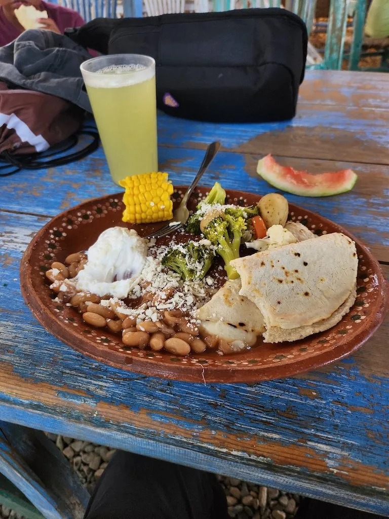 A brown cay plate with tortillas, vegetables, beans anc corn, and a glass of a yellow juice on a table.