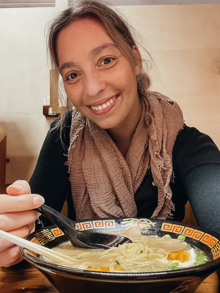 Woman with a black bowl of ramen with eggs smiling at the camera.