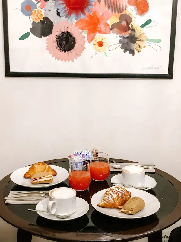 Image of a small round table with two mugs of coffee, two glasses of juice, and two plates with pastries and a sandwich