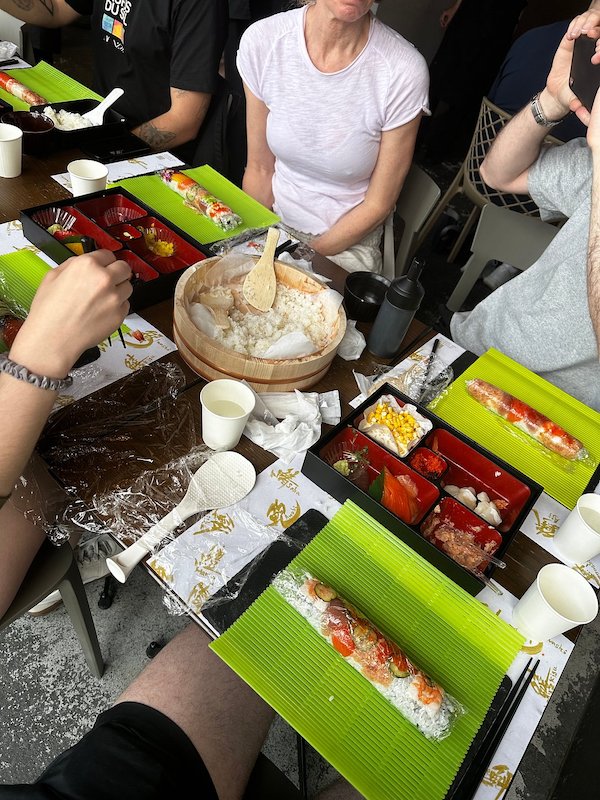 Multiple people during a cooking class around a table covered with rice and sushi mats with rolled sushi.