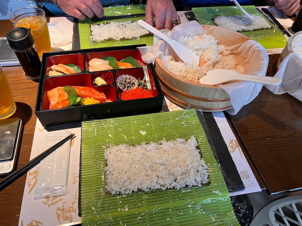 A green sushi mat covered wurg algea and rice, and in the background a bowl of white rice and a bento box with various type sof fish.