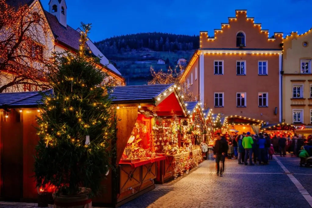 An outdoor square at night with Christmas Market stalls lit up in fairy lights, and buildings in the background