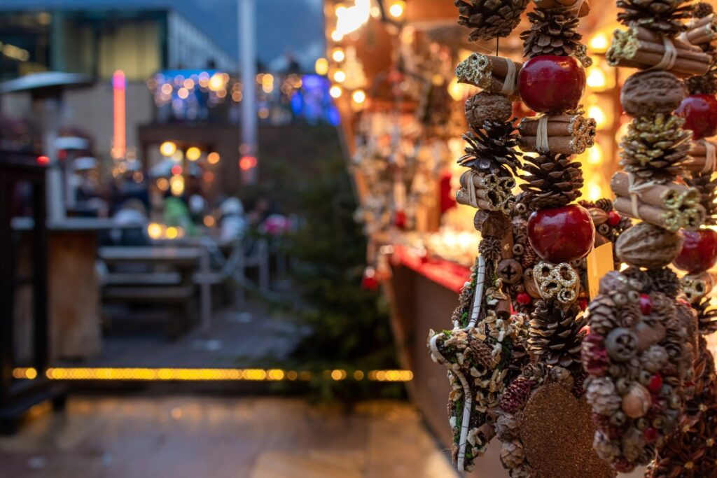 Christmas decorations made of small pine cones and apples seen from up close, and a blurry Christmas Market stall in the back