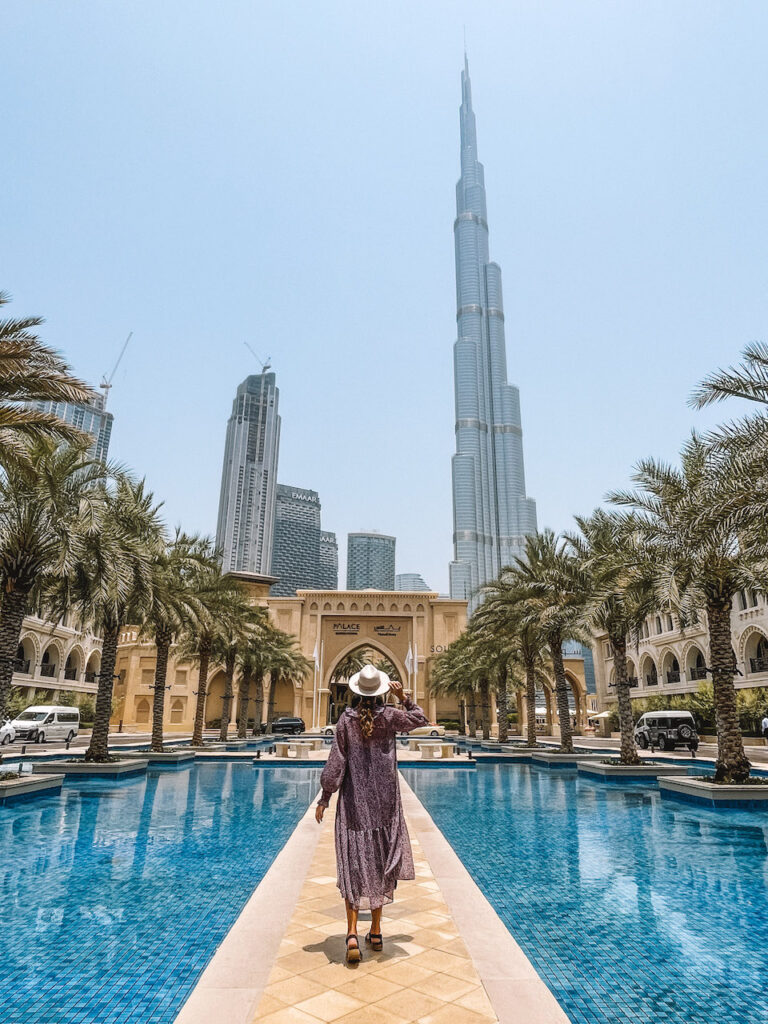 A woman looking up at the Burj Khalifa and two other skyscrapers as she stands in a walking board in the middle of a pool, lined with palm trees 