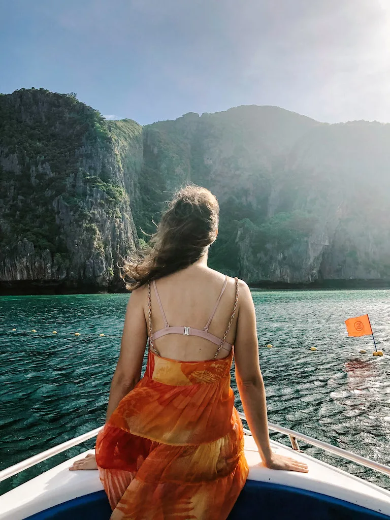 A woman from the back, wearing an orange sundress is standing at the front of a boat with Phang Nga bay rock formations in the background.