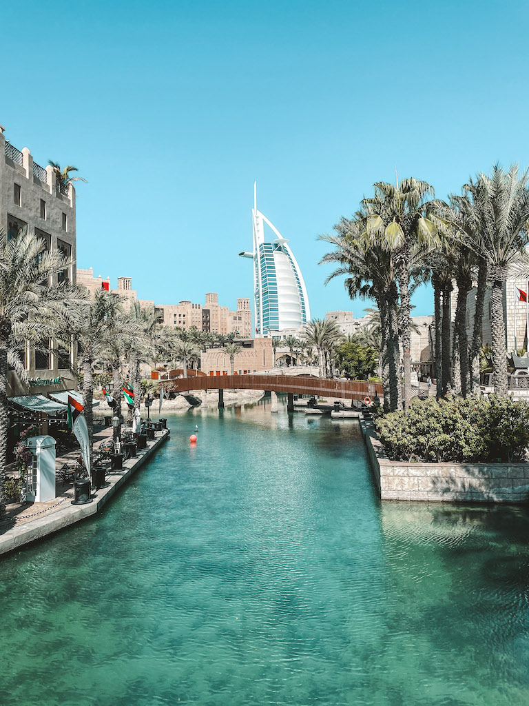 AN image of the Burj Al Arab in the distance, and the buildings of Souk Madinat Jumeirah, palm trees, and a canal in the forefront 