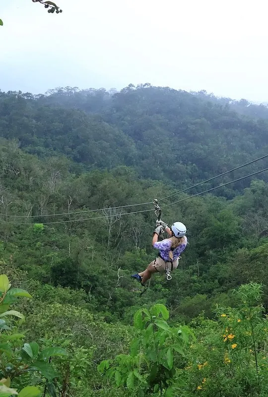 A woman wearing a while helmet is ziplining through dense jungle in Puerto Vallarta.