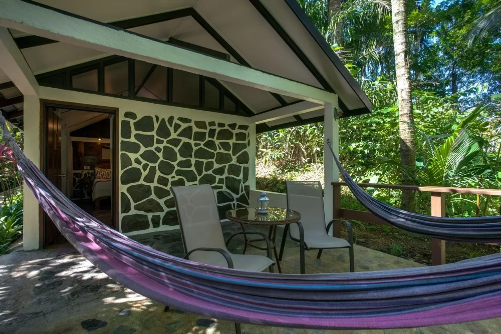 Two purple hammocks hanging on a terrace, with two chairs on both sides of a small round table, and a house in the back