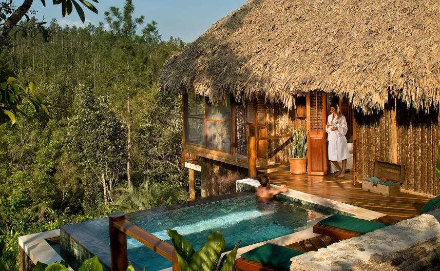 A woman in a white bathrobe leaning against a door on a thatched-roof wooden cabin, talking to a man in a swimming pool in front of her