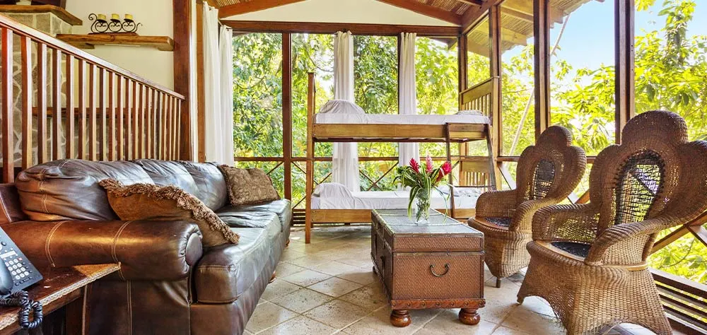 The interior of a lodge with a large brown sofa, two individual chairs and bunk beds in the back, with floor-to-ceiling glass windows on two sides