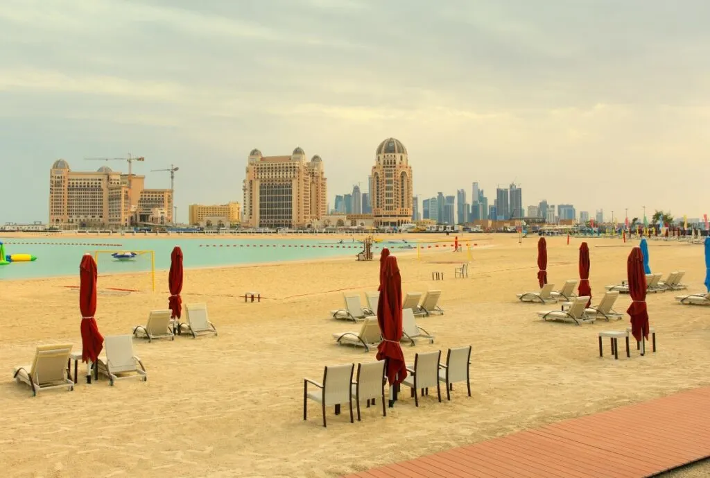 Katara beach, with sunbeds and chairs on the sun, closed red umbrellas, and buildings in the distance