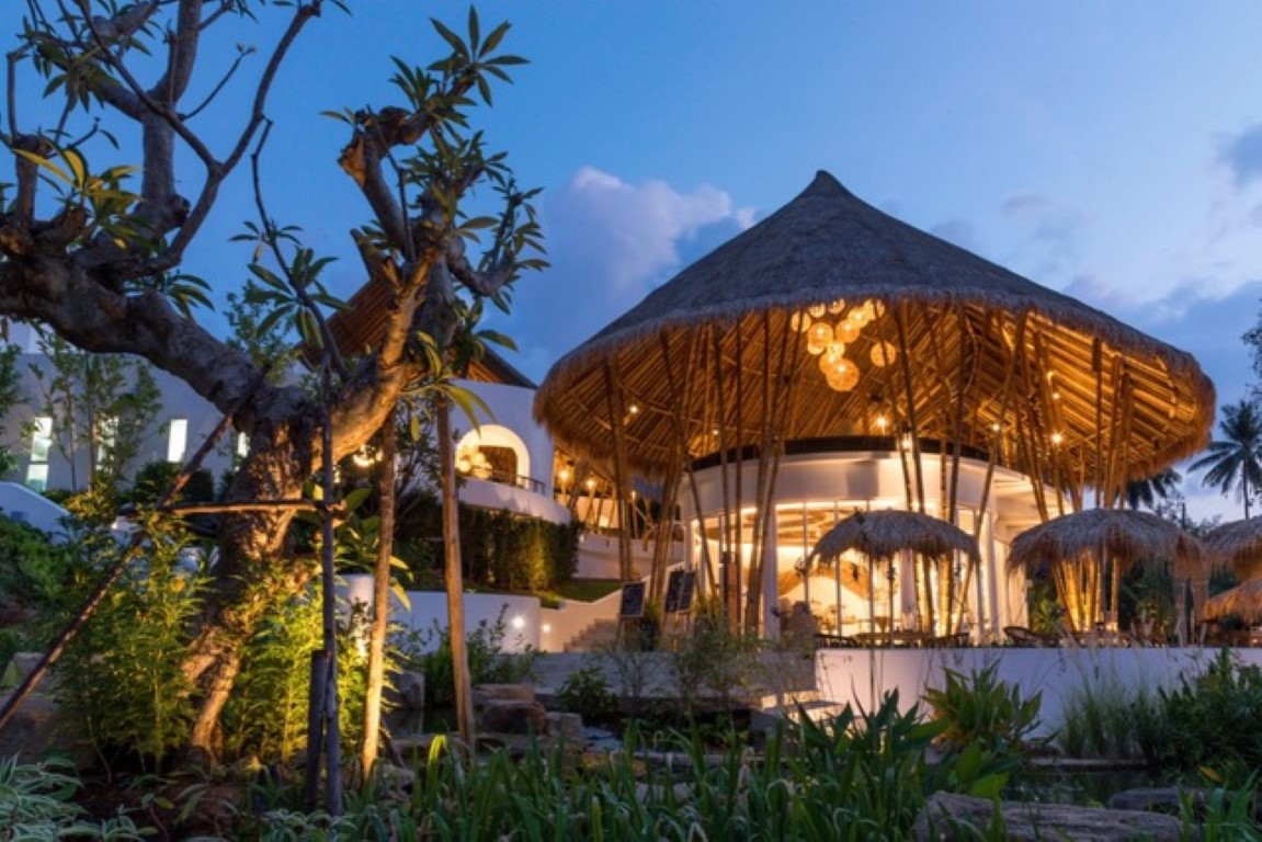 The entrance of a hotel at dusk with a thatched-roofed hall and plants 