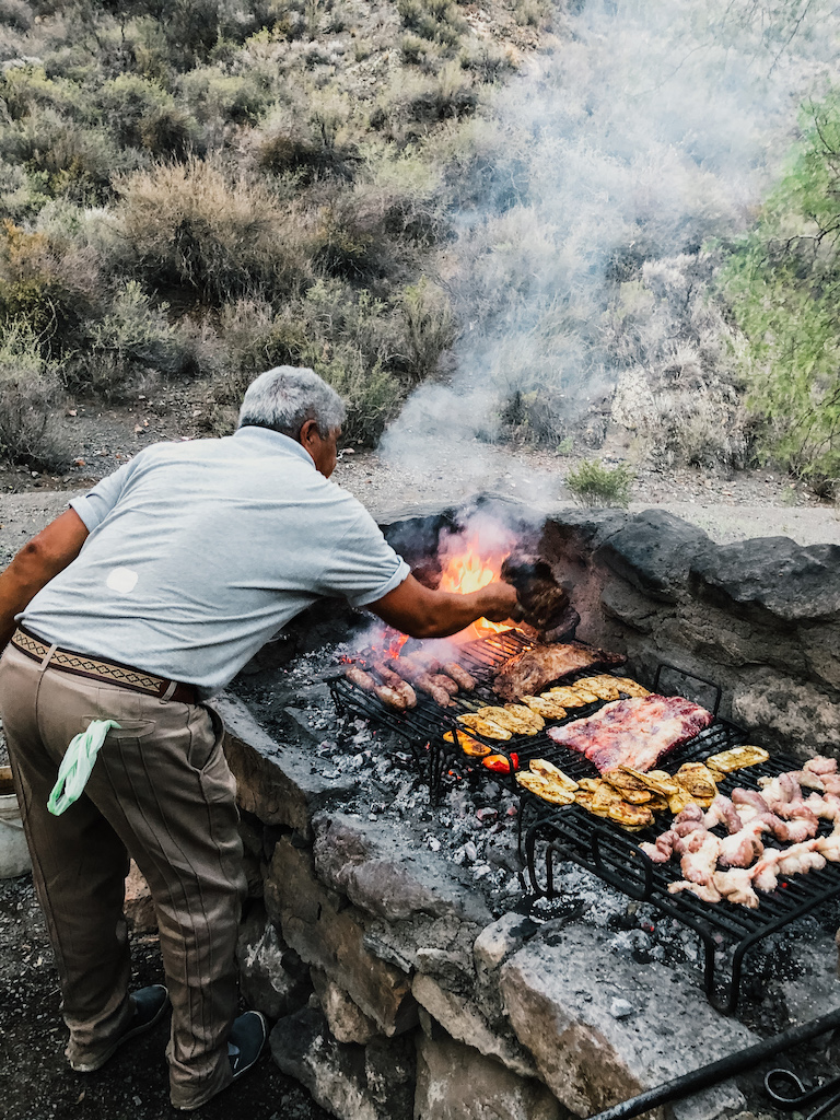 A traditional asado (barbecue) being prepared on a stone grill in Mendoza, Argentina, with various cuts of meat cooking over open flames.