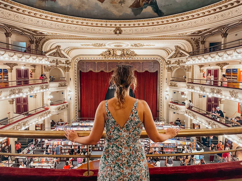 A woman standing in an indoor balcony overlooking a massive library below 