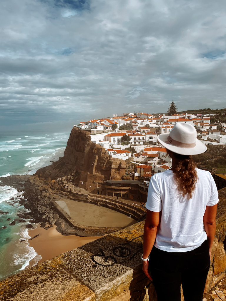 A woman in a white t-shirt, black trousers, and a hat overlooking a white town with terracotta roofs, and the beach 