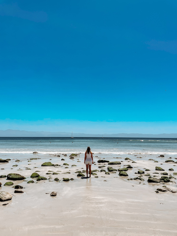 A woman standing on the sand of a beach, with rocks dotting the shoreline