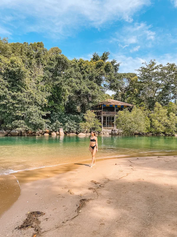 A woman walking along a beach, with lush vegetation in the background 