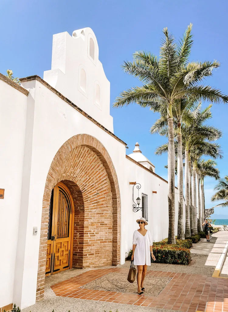 A woman in a white dress standing outside a colonial  building, with palm trees behind her