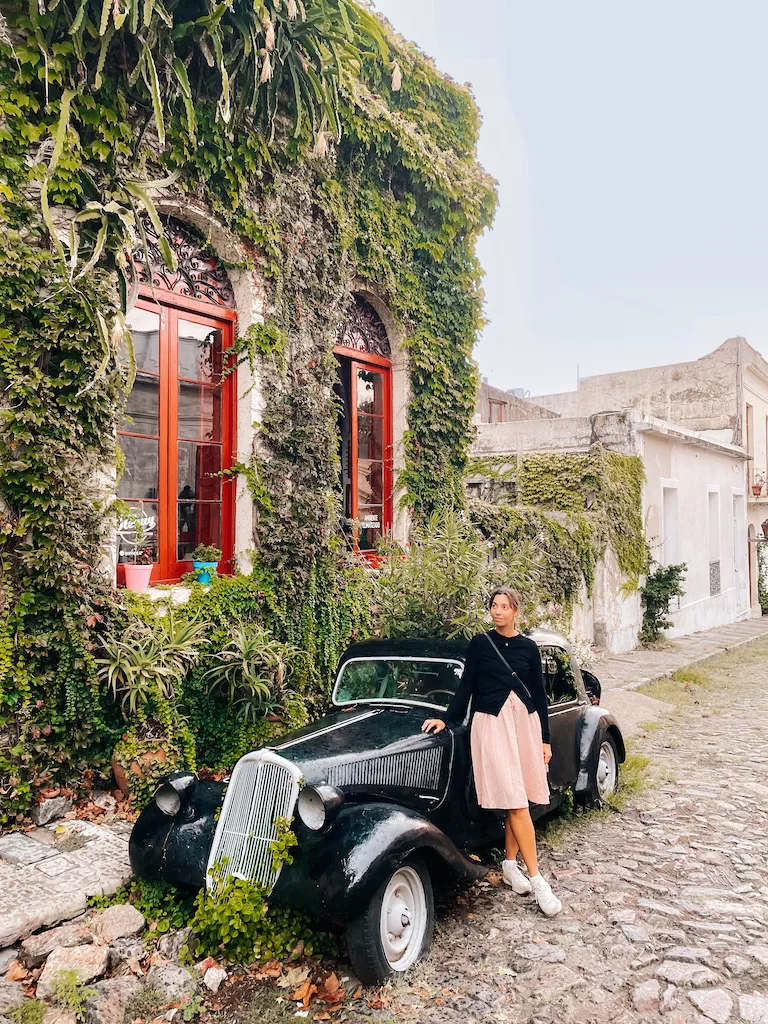 A woman standing next to a classic black car parked outside a building covered in vegetation 