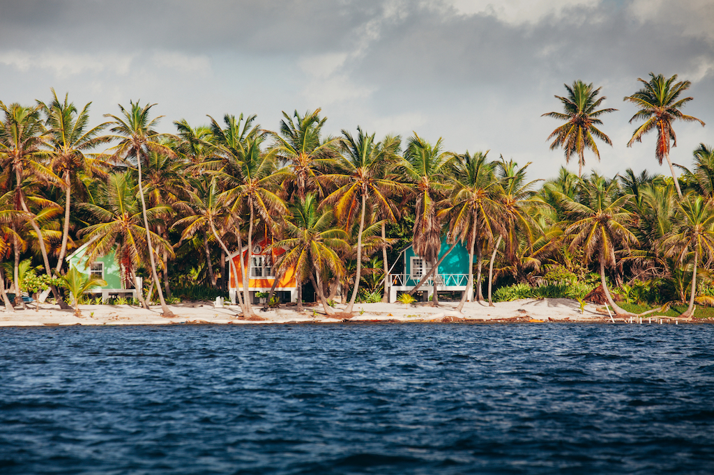 Colorful beachfront cabins surrounded by lush palm trees on a sandy shore in Belize, with the turquoise Caribbean Sea in the foreground.