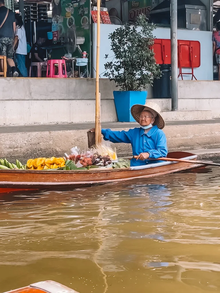 Vendor in traditional hat and blue shirt selling fresh fruit and snacks from a wooden boat in a floating market in Bangkok.