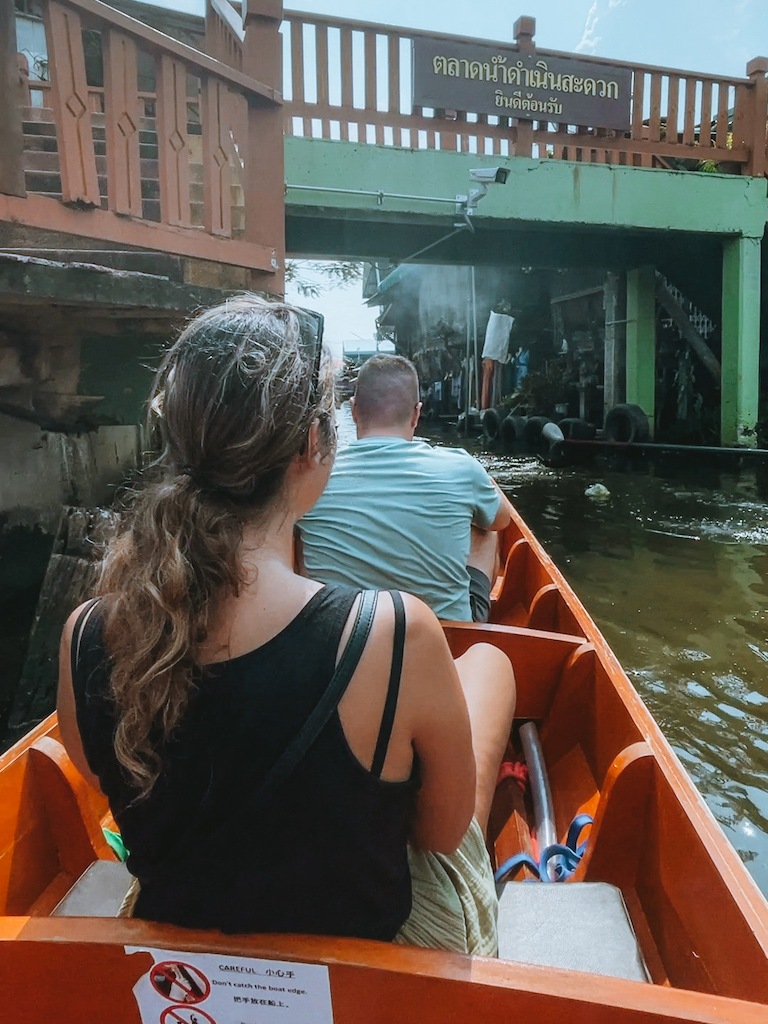 Two tourists riding in an orange wooden boat through Bangkok's floating market canals.