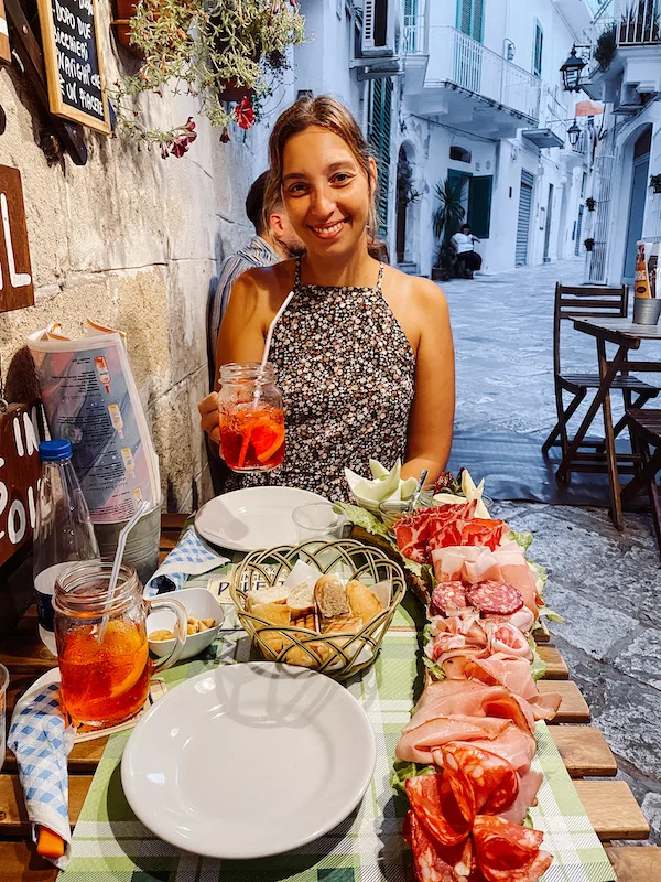 A woman smiling and holding an aperol spritz, in front of a table with a charcuterie board and a basket of bread