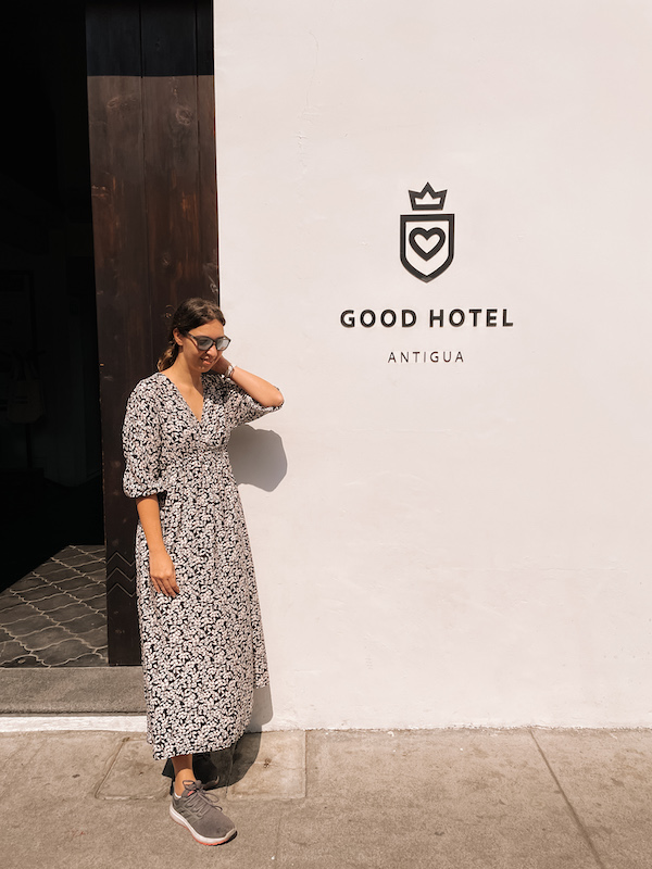 A woman in a long dress standing next to a wall that reads ''Good Hotel Antigua''