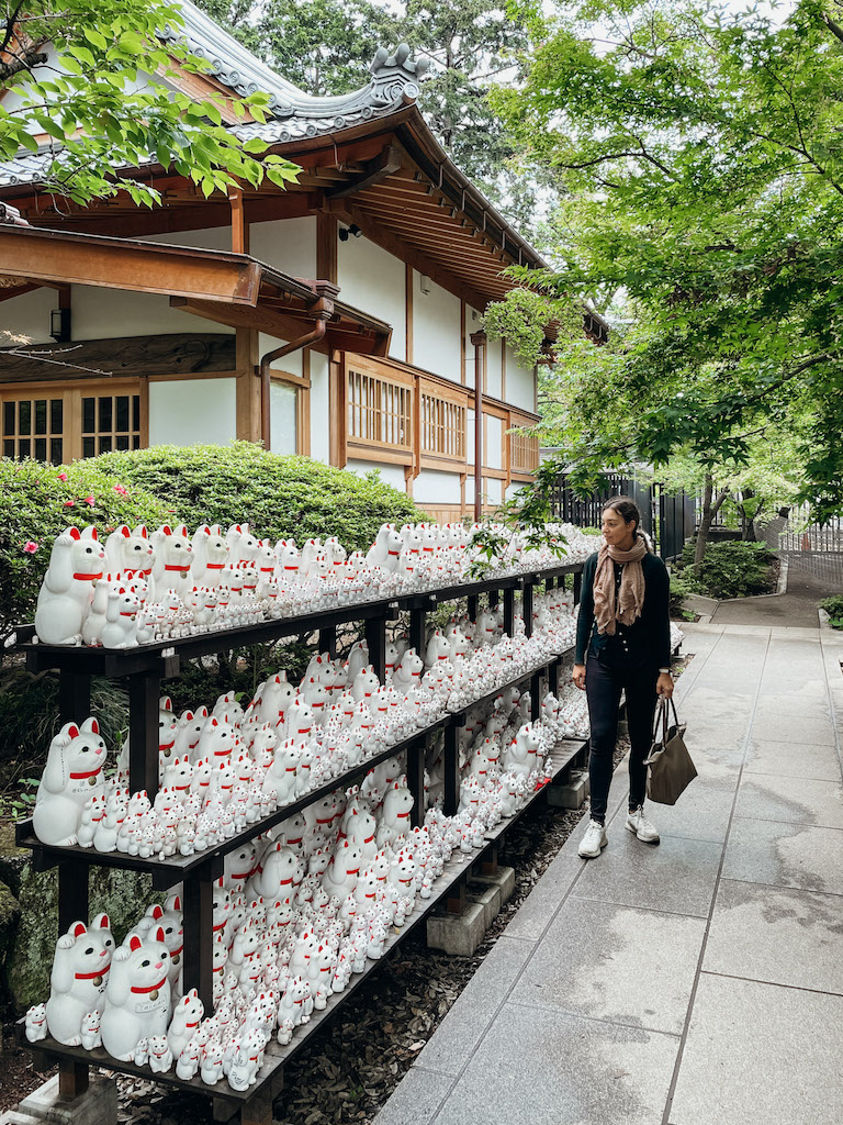 A woman standing next to a black shelf full of  Maneki-Neko cat figurines of various sizes outside a temple