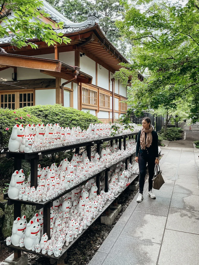 A woman standing in front of a long set of shelves with maneki neko cat figurines