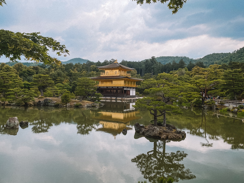 Photo of the golden Kinkaku-ji temple in Kyoto from the otehr side of a lake The temple is surrounded by greenery and is reflected in the water.