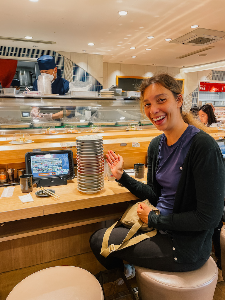 A woman smiling, sitting next to a sushi belt and a tower of small plates  